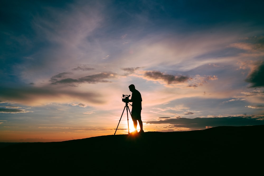 silhouette of man standing beside camera tripod during sunset