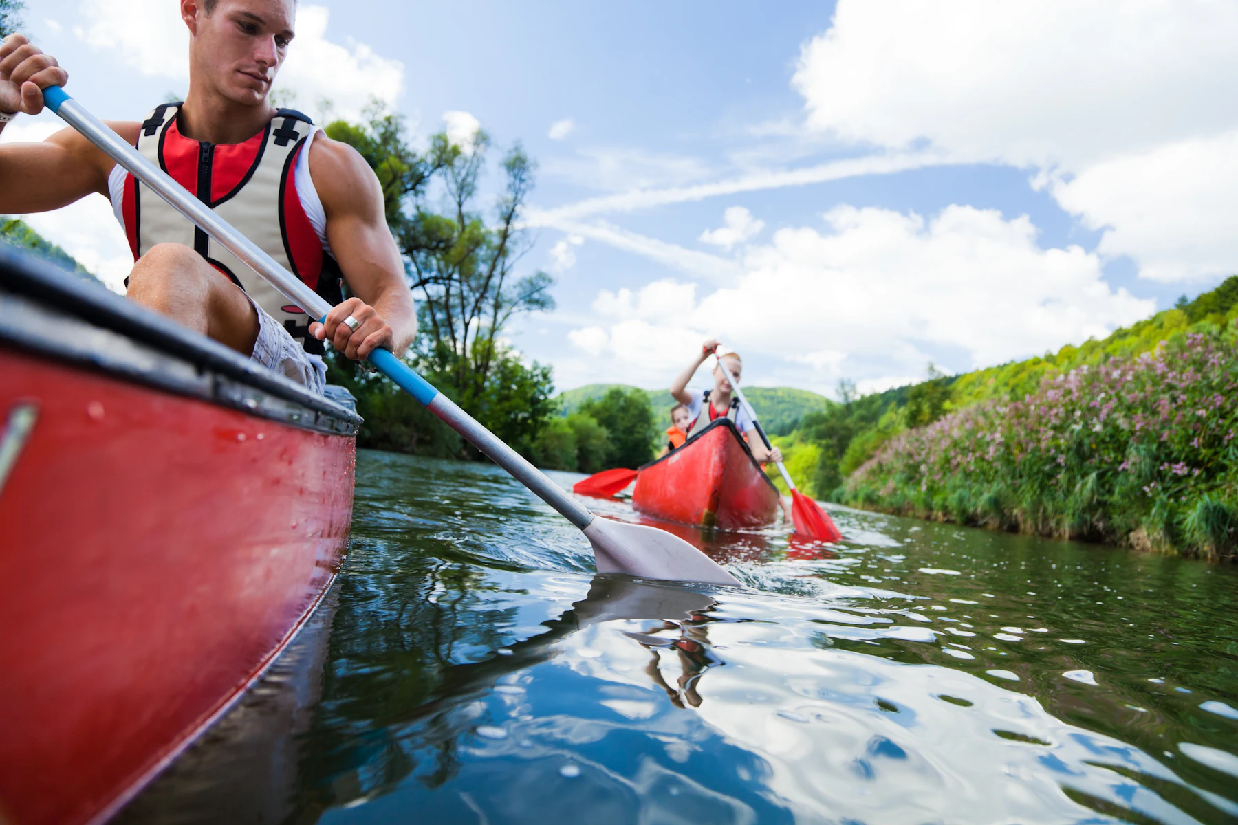 Two red canoes paddling a green river, seen low from the lead boat