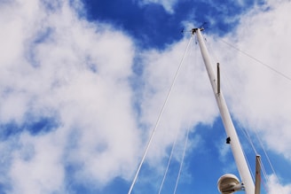 A tall sailboat mast extends into a sky filled with patches of white, fluffy clouds. Several ropes are attached to the mast, which also holds equipment such as an anemometer and a radar dome.