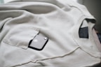A dark smartphone is partly visible in the pocket of a light beige or off-white sweatshirt. The sweatshirt is laid out flat, with its seams and pocket details clearly visible.