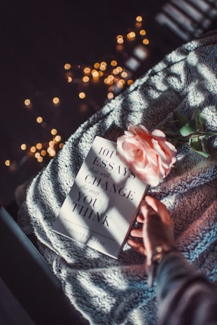 A book titled '101 Essays that will Change the Way You Think' lies on a soft, textured blanket. A pale pink rose is placed on the book, and a hand is lightly resting on the corner. Soft, warm fairy lights glow in the background, creating a cozy atmosphere.