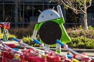 A large Android figurine is holding an Oreo cookie. The scene includes colorful bicycles with red baskets in the foreground. The background features a building with glass windows and landscaped greenery, including a small tree.