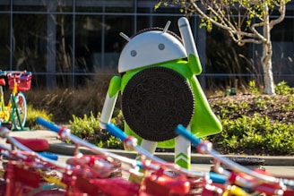 A large Android figurine is holding an Oreo cookie. The scene includes colorful bicycles with red baskets in the foreground. The background features a building with glass windows and landscaped greenery, including a small tree.