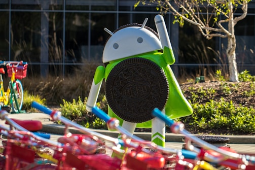 A large Android figurine is holding an Oreo cookie. The scene includes colorful bicycles with red baskets in the foreground. The background features a building with glass windows and landscaped greenery, including a small tree.