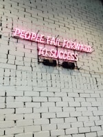 A pink neon sign with the text 'PEOPLE FAIL FORWARD TO SUCCESS' mounted on a white brick wall. The bricks are uniform in size and the wall is clean and well-maintained.