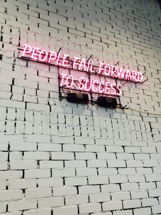 A pink neon sign with the text 'PEOPLE FAIL FORWARD TO SUCCESS' mounted on a white brick wall. The bricks are uniform in size and the wall is clean and well-maintained.