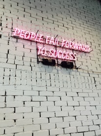A pink neon sign with the text 'PEOPLE FAIL FORWARD TO SUCCESS' mounted on a white brick wall. The bricks are uniform in size and the wall is clean and well-maintained.
