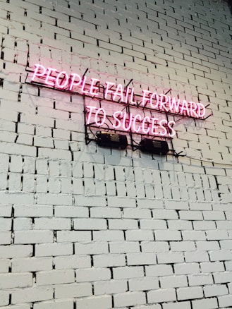 A pink neon sign with the text 'PEOPLE FAIL FORWARD TO SUCCESS' mounted on a white brick wall. The bricks are uniform in size and the wall is clean and well-maintained.