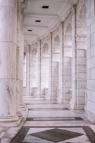 A curved corridor with large marble columns and a polished stone floor. The architectural design features arched openings and a high ceiling, creating a sense of elegance and grandeur.