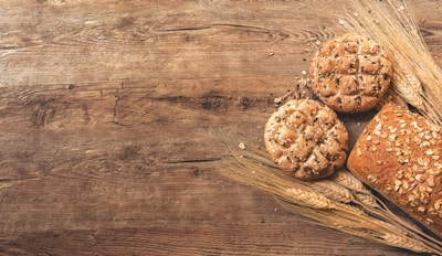 Several pieces of rustic bread with seeds and grains are placed on a wooden surface. The breads are surrounded by stalks of wheat, creating a natural and rustic composition.