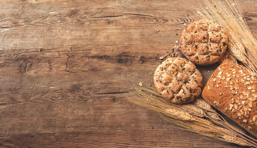 Several pieces of rustic bread with seeds and grains are placed on a wooden surface. The breads are surrounded by stalks of wheat, creating a natural and rustic composition.