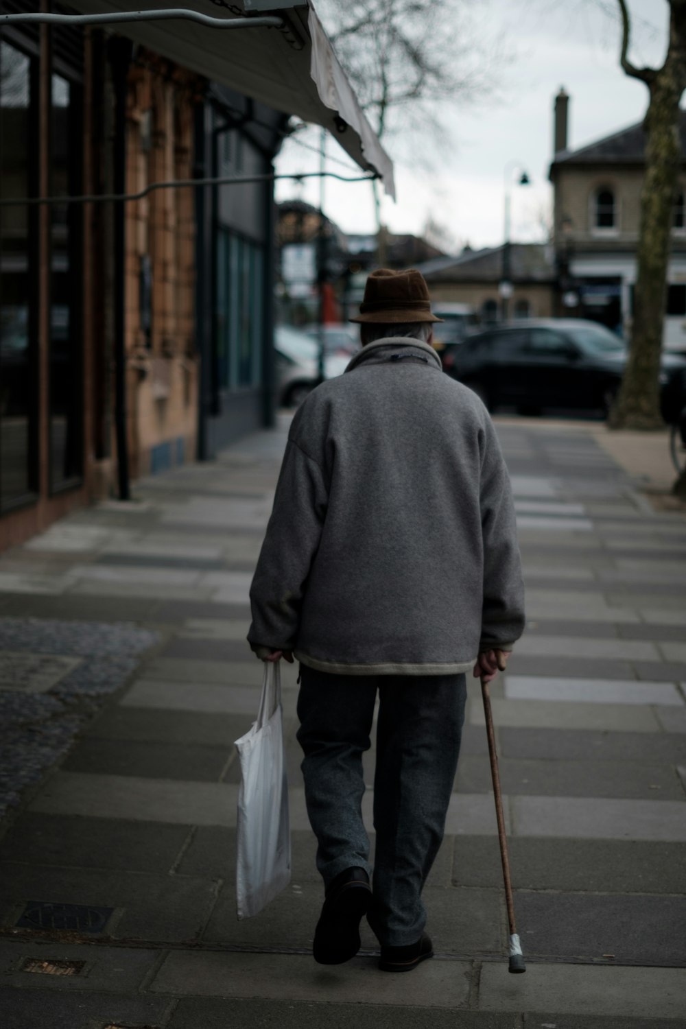 昼間、茶色の杖をついて白い袋を持って茶色の建物の近くを歩く男性