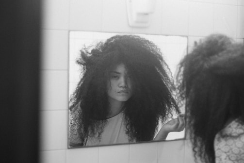 A person with voluminous, curly hair is looking at themselves in a bathroom mirror. The image is in black and white, and the setting is simple with tiled walls. The person has a contemplative expression, and their hand is gently touching their hair.