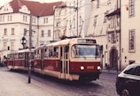A vintage red and cream tram travels through a European city street lined with historic buildings featuring tall windows and intricate architectural details. Overhead power lines and a classic street clock add to the old-world charm. The cobblestone street has parked cars and the setting feels quiet and nostalgic.