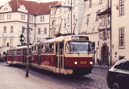 A vintage red and cream tram travels through a European city street lined with historic buildings featuring tall windows and intricate architectural details. Overhead power lines and a classic street clock add to the old-world charm. The cobblestone street has parked cars and the setting feels quiet and nostalgic.