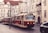 A vintage red and cream tram travels through a European city street lined with historic buildings featuring tall windows and intricate architectural details. Overhead power lines and a classic street clock add to the old-world charm. The cobblestone street has parked cars and the setting feels quiet and nostalgic.