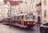 A vintage red and cream tram travels through a European city street lined with historic buildings featuring tall windows and intricate architectural details. Overhead power lines and a classic street clock add to the old-world charm. The cobblestone street has parked cars and the setting feels quiet and nostalgic.