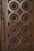 A wooden door with intricate circular and diamond-shaped carvings arranged in a symmetrical pattern. The design is rich and ornate, with dark brown tones and a metallic handle on the left side.