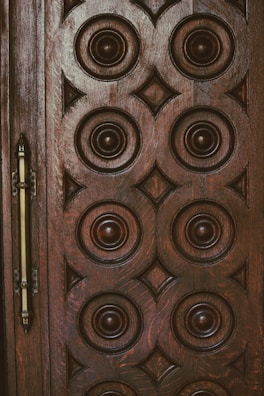 A wooden door with intricate circular and diamond-shaped carvings arranged in a symmetrical pattern. The design is rich and ornate, with dark brown tones and a metallic handle on the left side.
