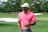 A person wearing a pink polo shirt and white cap stands on a golf course holding a golf glove. The background shows a green fairway with sand traps and trees.