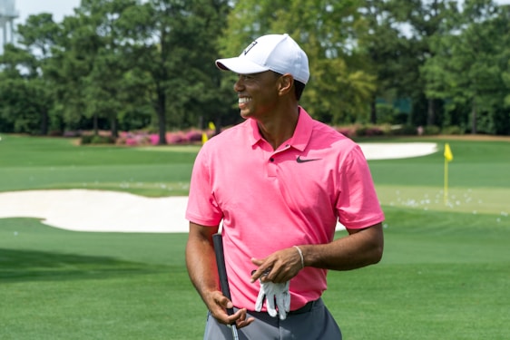 A person wearing a pink polo shirt and white cap stands on a golf course holding a golf glove. The background shows a green fairway with sand traps and trees.
