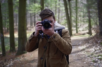 A person wearing a brown jacket and holding a Canon camera is taking a photograph in a wooded area. The background is filled with tall green trees and a dirt path.
