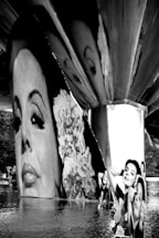 A black and white image features large photographic panels under a bridge. The main focus is on portraits of a woman's face, partially submerged in water, reflecting a serene and contemplative expression. The panels create an artistic installation that blends with the calm surface of the water, capturing a unique juxtaposition between the industrial bridge structure and the human face imagery.
