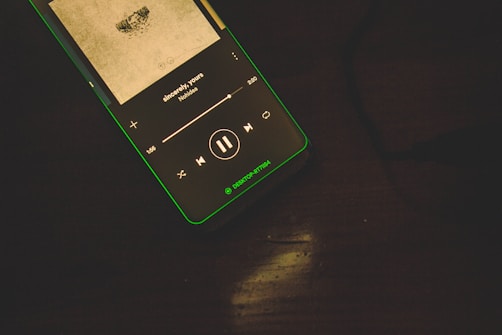 A smartphone lays on a dark surface displaying a music player interface. The screen shows album art and song information, including the title 'sincerely yours' by an artist named No1dea. The interface features music control icons like play, pause, and track progression slider.
