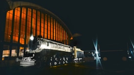 A vintage steam locomotive is parked in front of a large, arched, glass-fronted building illuminated with a warm, orange glow. The scene is set at night, and the darkness contrasts with the bright light from the train's headlamp and the building's interior.