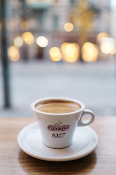 A white coffee cup with the text 'Carraro 1927' on it is placed on a saucer on a wooden table. In the background, there is a blurred view of lights and a cityscape, creating a bokeh effect.