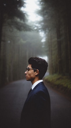 A person in a suit stands in profile on a foggy, forested road. Tall trees line the path, creating a serene yet mysterious atmosphere.