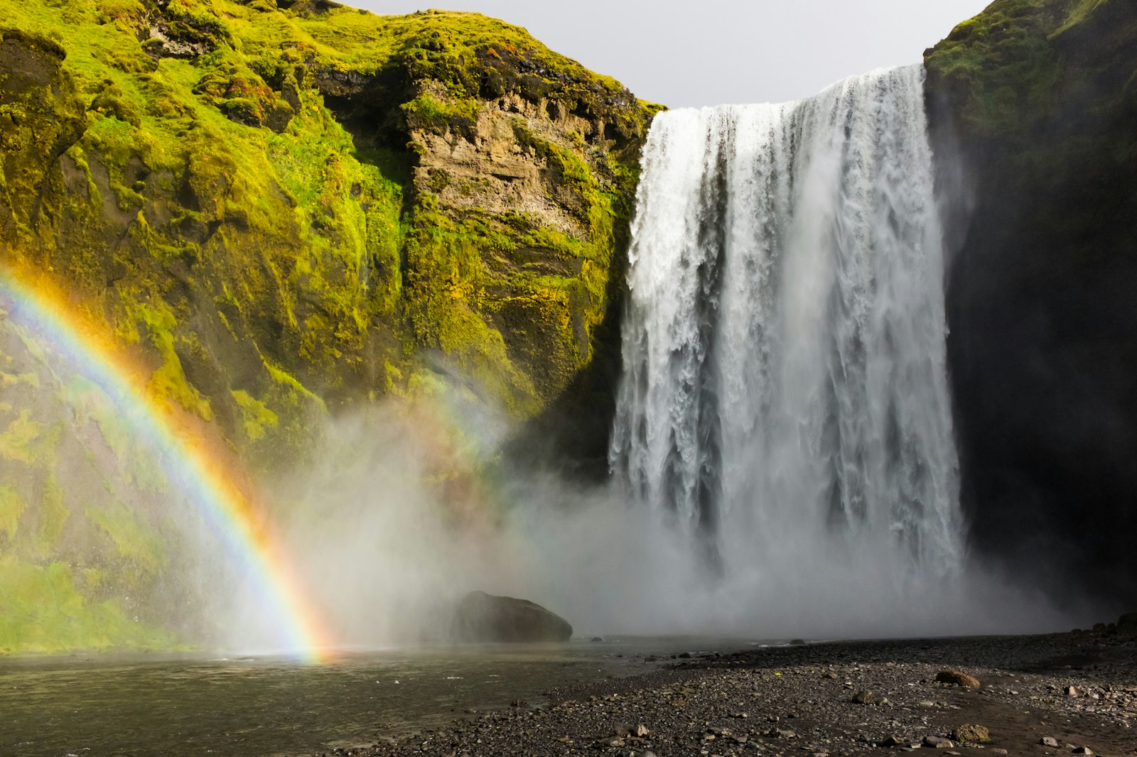A wide waterfall plunging off a green cliff with a rainbow forming in the spray near its base