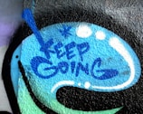 The image depicts a graffiti-style artwork on a textured surface. The words 'KEEP GOING' are spray-painted in blue within a speech bubble-like shape. Surrounding the text are abstract blue and white dots and lines, which add to the dynamic and encouraging visual appeal.