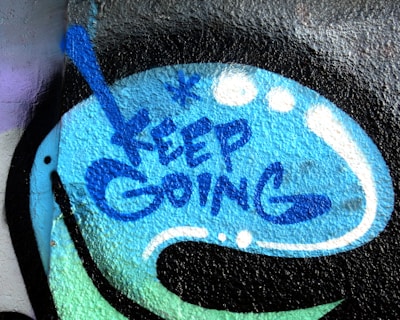 The image depicts a graffiti-style artwork on a textured surface. The words 'KEEP GOING' are spray-painted in blue within a speech bubble-like shape. Surrounding the text are abstract blue and white dots and lines, which add to the dynamic and encouraging visual appeal.