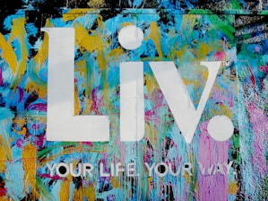 The image features a vibrant graffiti-style background with splashes of blue, yellow, pink, and green colors. Superimposed on this colorful backdrop are the large white letters 'Liv.' and the phrase 'YOUR LIFE YOUR WAY.' in additional white text.