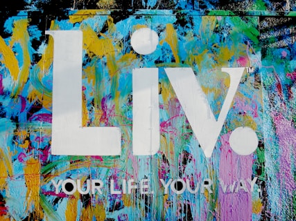 The image features a vibrant graffiti-style background with splashes of blue, yellow, pink, and green colors. Superimposed on this colorful backdrop are the large white letters 'Liv.' and the phrase 'YOUR LIFE YOUR WAY.' in additional white text.