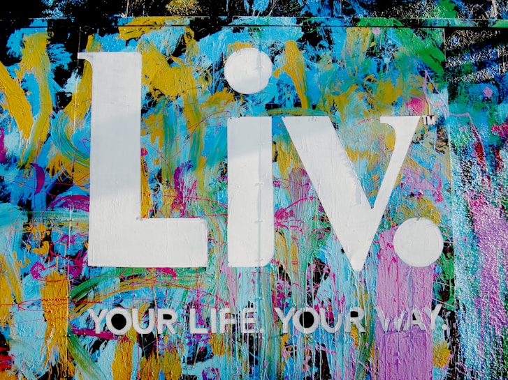 The image features a vibrant graffiti-style background with splashes of blue, yellow, pink, and green colors. Superimposed on this colorful backdrop are the large white letters 'Liv.' and the phrase 'YOUR LIFE YOUR WAY.' in additional white text.