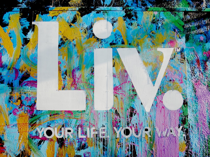 The image features a vibrant graffiti-style background with splashes of blue, yellow, pink, and green colors. Superimposed on this colorful backdrop are the large white letters 'Liv.' and the phrase 'YOUR LIFE YOUR WAY.' in additional white text.