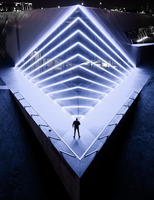 A person stands on a triangular concrete platform with bright neon blue lights creating geometric patterns along the edges. The scene is set at night, and the structure appears to be a modern architectural building with reflective surfaces. The overall ambiance is futuristic and atmospheric.