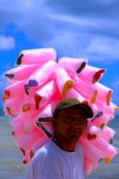 A person is carrying numerous bags of pink cotton candy on their back while standing near a body of water. The sky is clear with a few clouds, creating a vibrant and lively atmosphere.