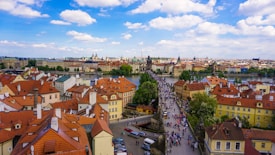 A scenic view of a European city with a river and a historic bridge filled with people. The foreground features buildings with red roofs, while the background shows more historical structures and a skyline under a partly cloudy blue sky.