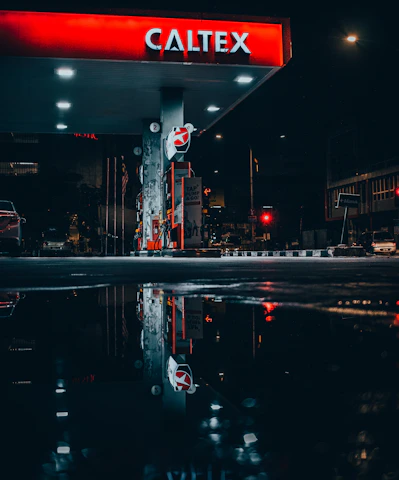 A gas station is illuminated at night, featuring a prominent red and white Caltex sign. The station's bright lights create reflections on the wet pavement below. The surrounding environment is dark, enhancing the contrast with the station's lights. Some cars and streetlights are visible in the background.