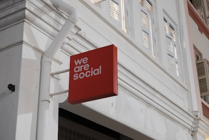 A red rectangular sign with the words 'we are social' is affixed to the exterior of a white building. The building features architectural elements such as window shutters and decorative molding, with visible piping attached to the wall.