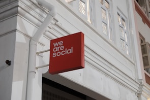 A red rectangular sign with the words 'we are social' is affixed to the exterior of a white building. The building features architectural elements such as window shutters and decorative molding, with visible piping attached to the wall.