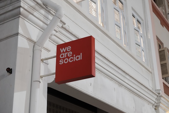 A red rectangular sign with the words 'we are social' is affixed to the exterior of a white building. The building features architectural elements such as window shutters and decorative molding, with visible piping attached to the wall.