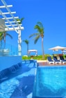 A luxurious outdoor swimming pool with cascading water and clear blue tiles, surrounded by palm trees. In the background, sun loungers with umbrellas and a gazebo can be seen on a wooden deck, creating a serene tropical atmosphere under a clear blue sky.