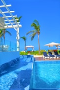 A luxurious outdoor swimming pool with cascading water and clear blue tiles, surrounded by palm trees. In the background, sun loungers with umbrellas and a gazebo can be seen on a wooden deck, creating a serene tropical atmosphere under a clear blue sky.