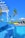 A luxurious outdoor swimming pool with cascading water and clear blue tiles, surrounded by palm trees. In the background, sun loungers with umbrellas and a gazebo can be seen on a wooden deck, creating a serene tropical atmosphere under a clear blue sky.