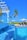 A luxurious outdoor swimming pool with cascading water and clear blue tiles, surrounded by palm trees. In the background, sun loungers with umbrellas and a gazebo can be seen on a wooden deck, creating a serene tropical atmosphere under a clear blue sky.