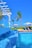 A luxurious outdoor swimming pool with cascading water and clear blue tiles, surrounded by palm trees. In the background, sun loungers with umbrellas and a gazebo can be seen on a wooden deck, creating a serene tropical atmosphere under a clear blue sky.